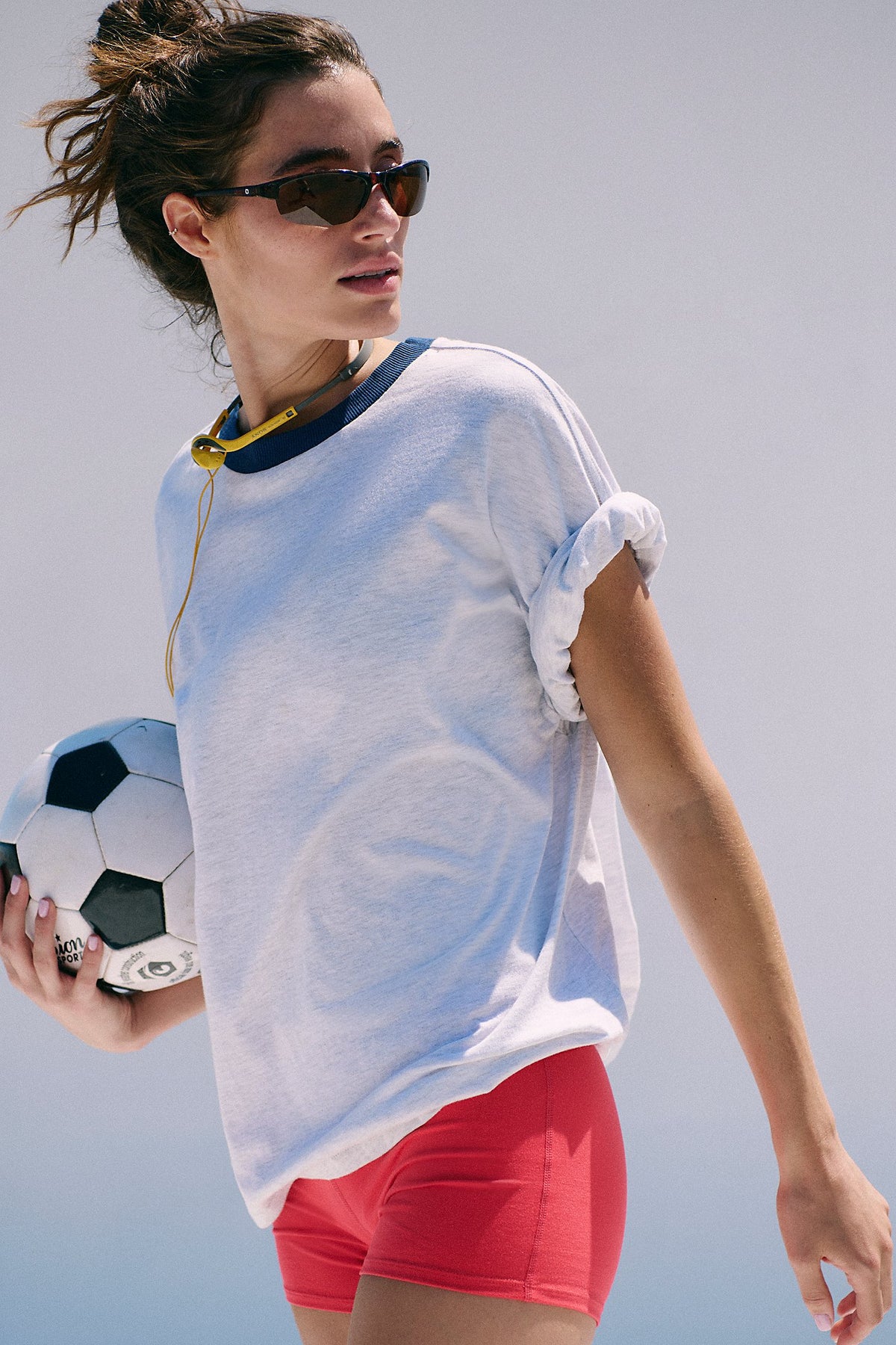 Woman holding a soccer ball wearing a white oversized shirt and red shorts against a light background