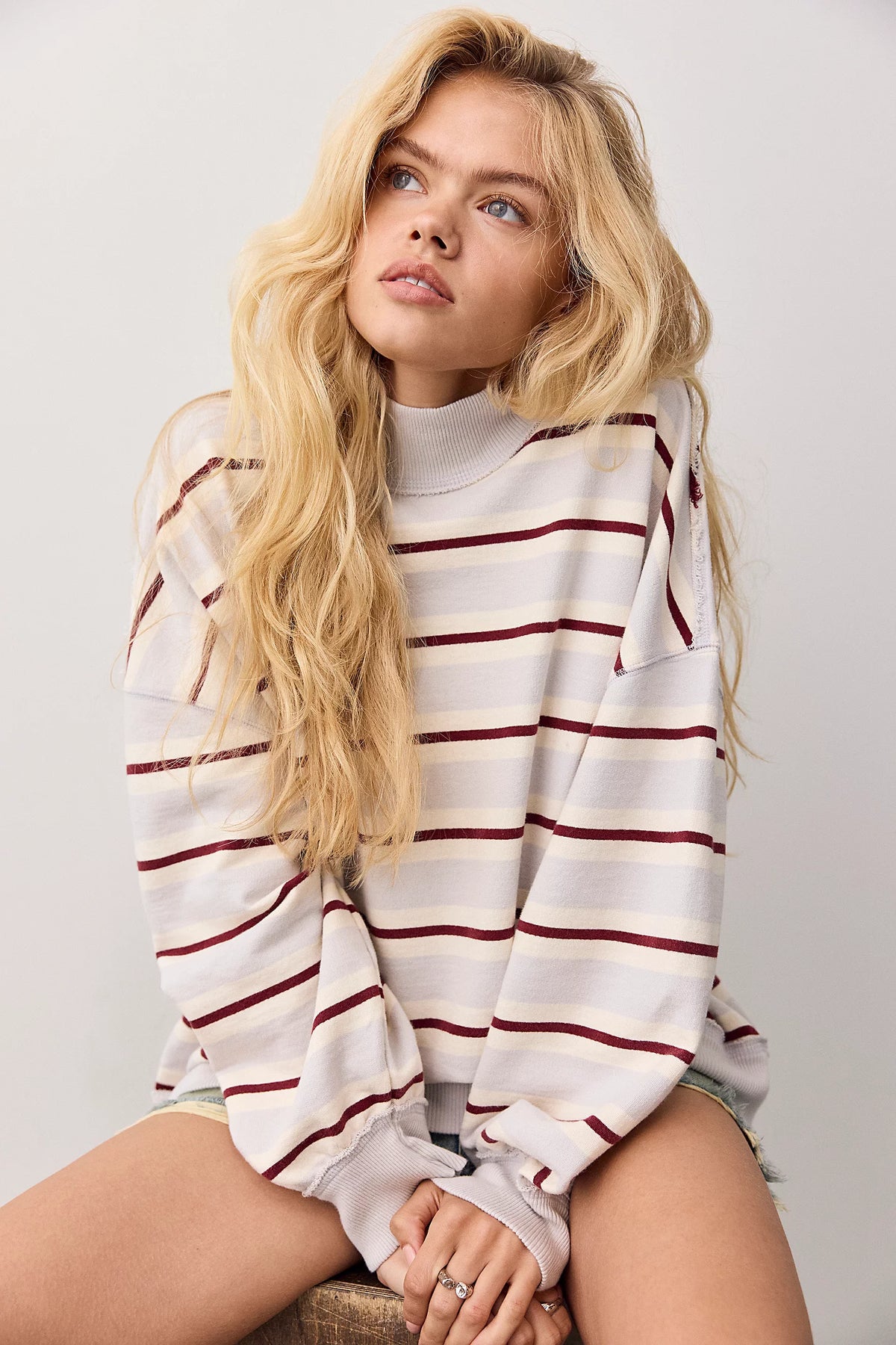 A blonde woman wearing a white long-sleeve mock neck sweatshirt with thin dark red horizontal stripes, sitting against a light gray background.