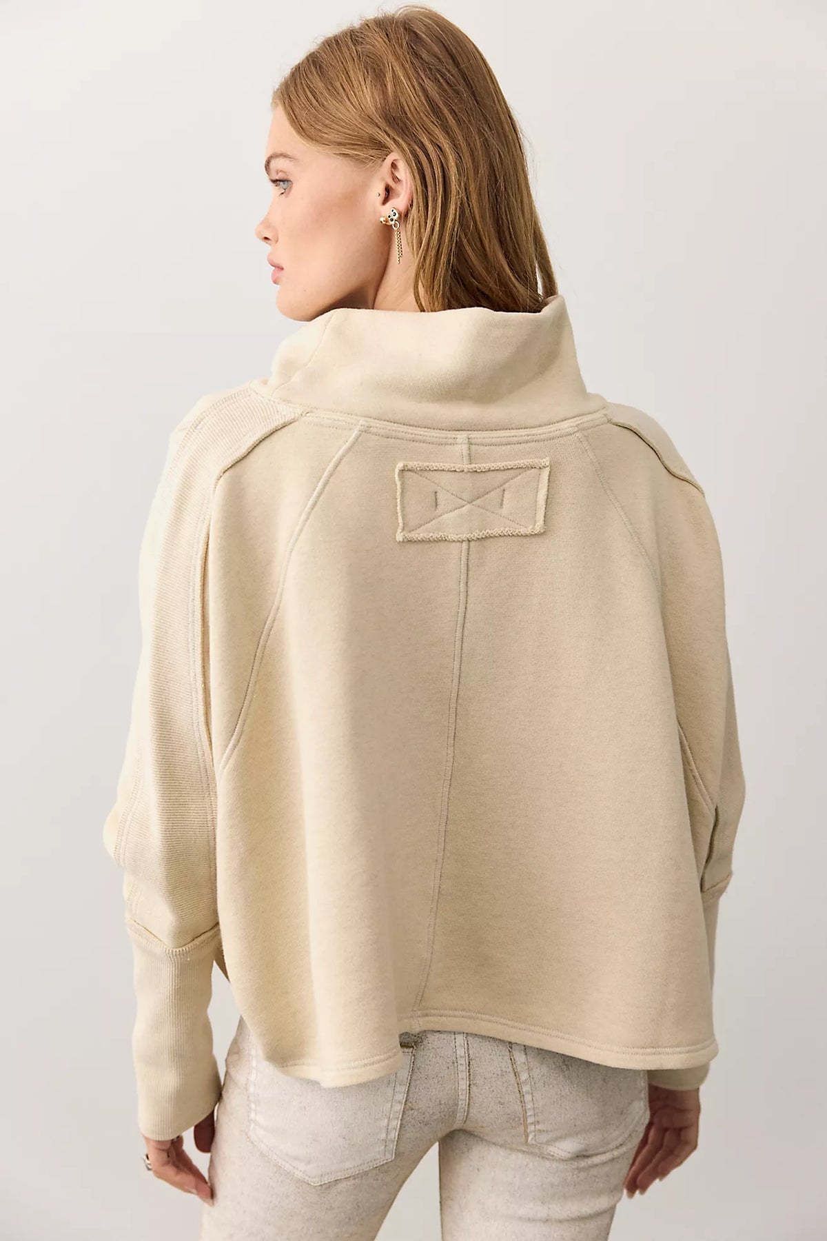 A woman shown from the back wearing a cropped beige mock-neck sweatshirt with shoulder patch details, paired with light gray jeans, against a plain light gray studio background.
