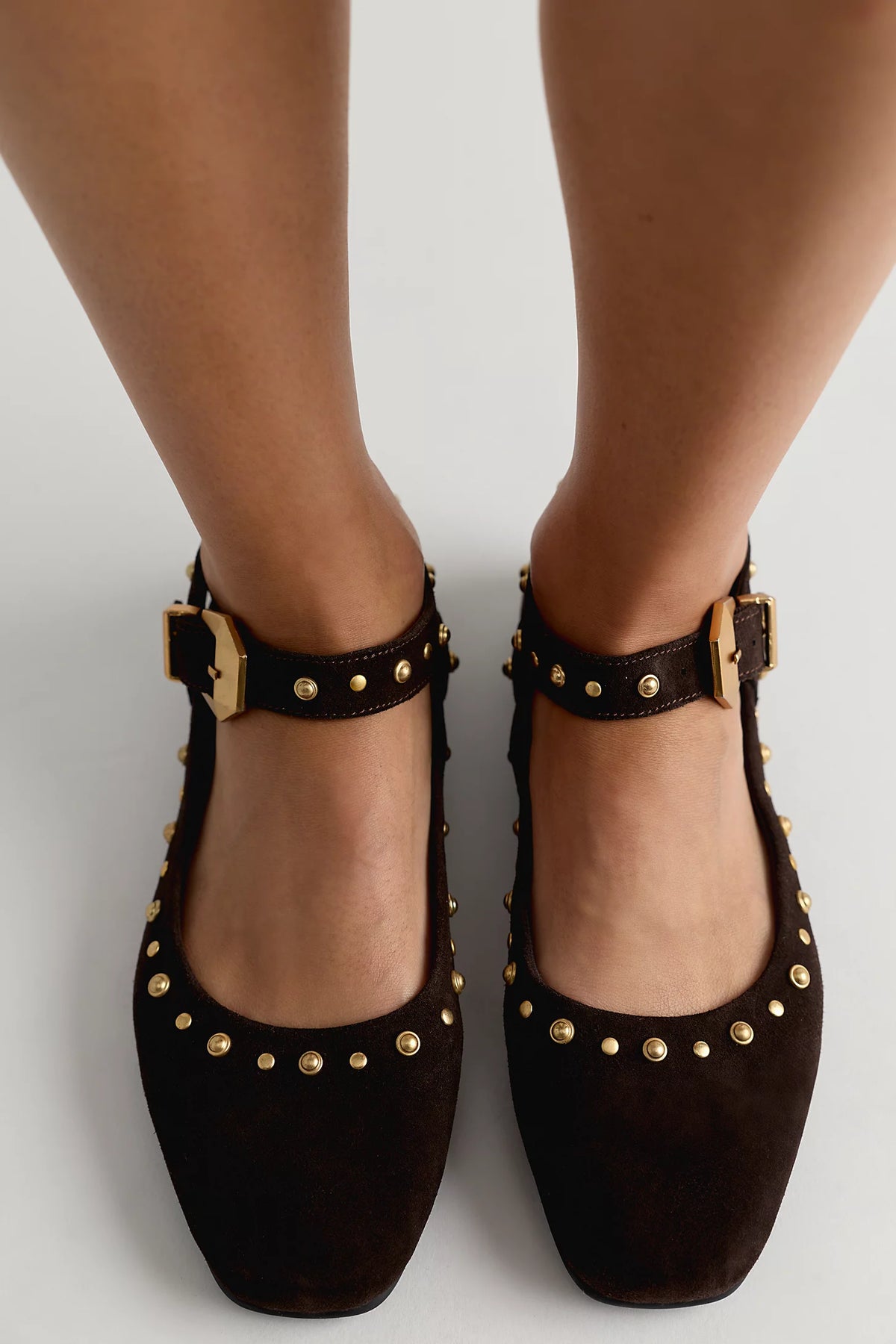 A pair of dark brown suede Mary Jane shoes with gold-tone studs and buckles, shown on a model's feet against a light gray background.