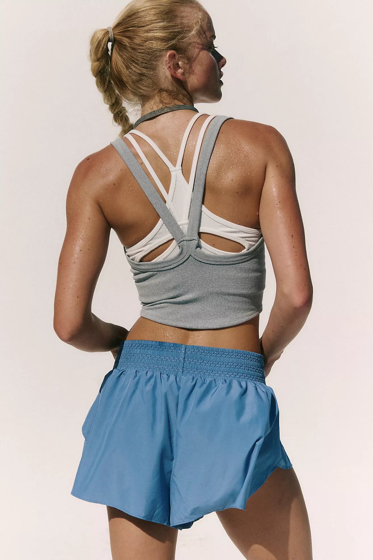 A woman shown from the back wearing a gray athletic crop top with white crisscross straps and a blue athletic skirt, against a plain light background.
