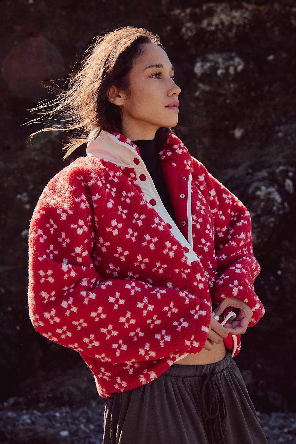 A woman wearing a red fuzzy cropped jacket with a white floral pattern and white zipper details, photographed outdoors against a dark rocky background.