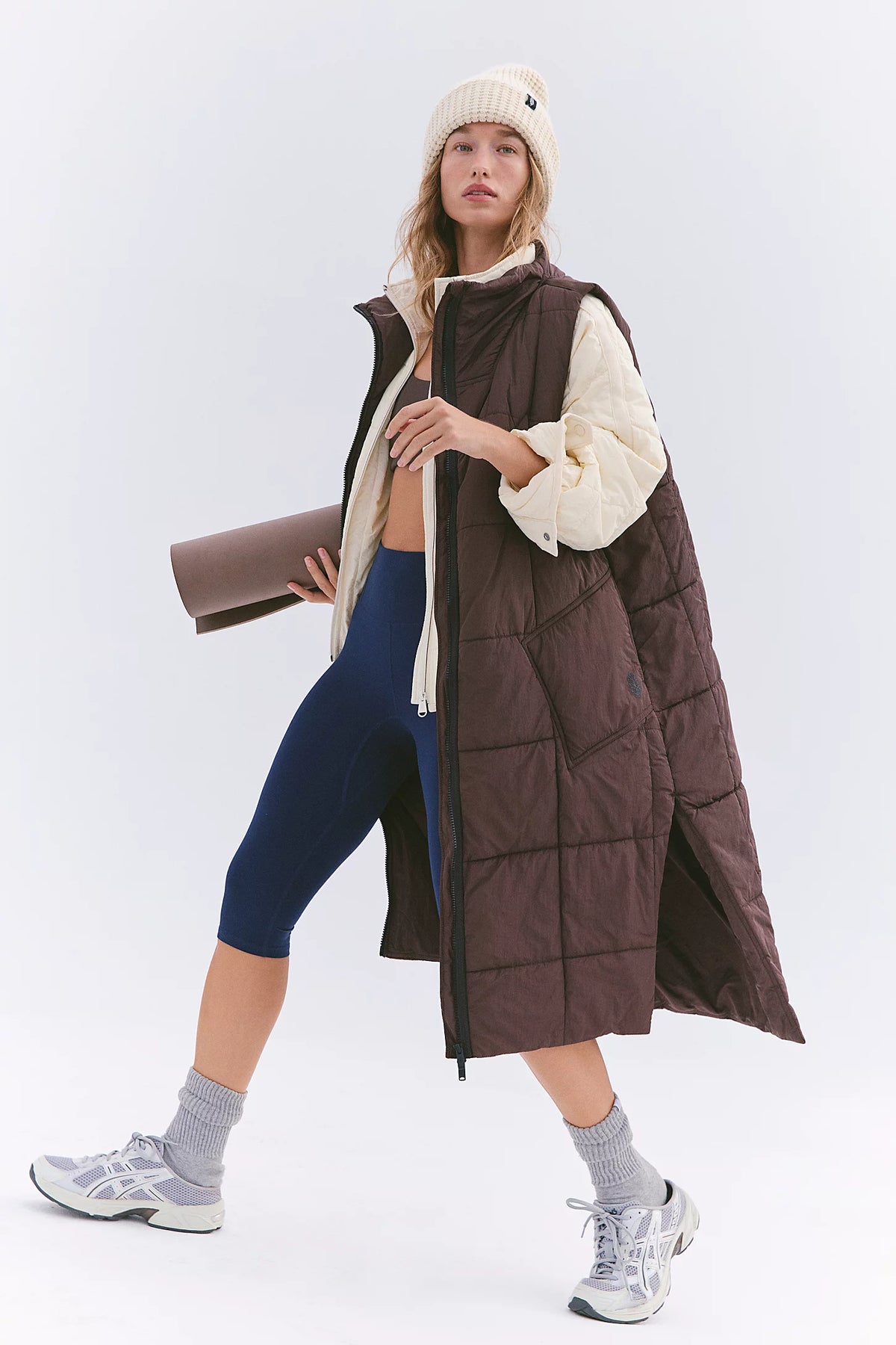 A woman models a long brown puffer vest with a cream sherpa lining over a cream zip-up hoodie, navy blue capri leggings, gray socks, and white sneakers against a light gray studio background.
