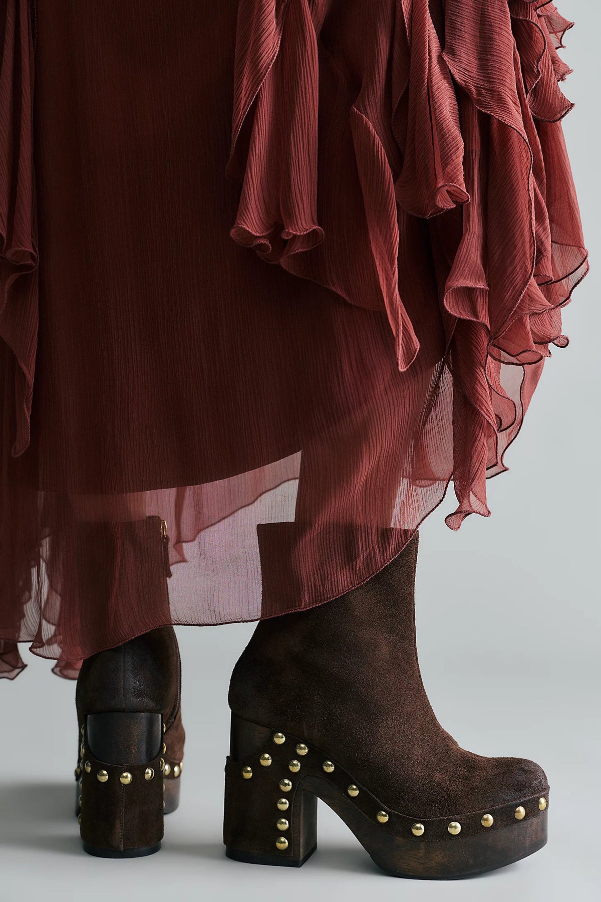 A close-up of a person wearing dark brown suede platform ankle boots with gold-tone studs, paired with a rust-colored sheer ruffled skirt, against a light gray studio background.