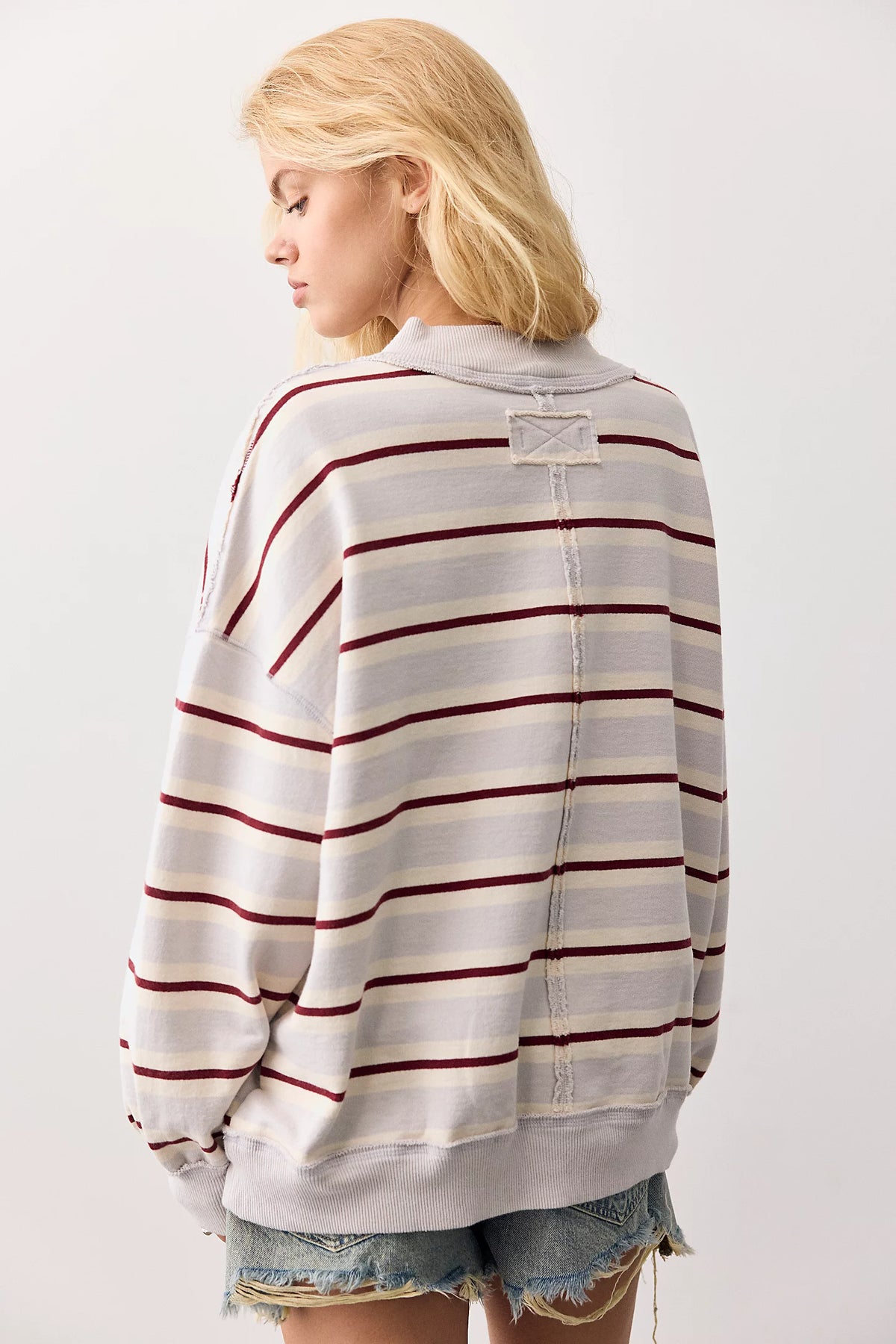 A woman shown from the back wearing an oversized striped sweatshirt with horizontal burgundy and cream stripes on a light gray base, paired with frayed denim shorts against a plain light gray studio background.