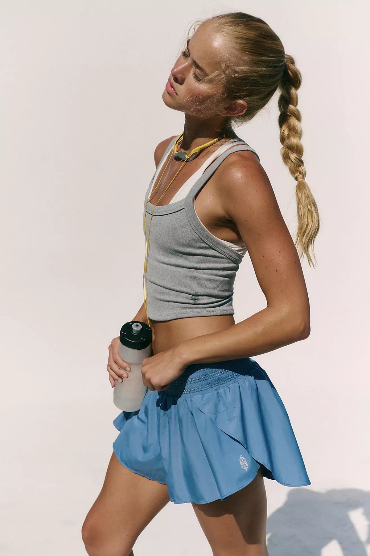 A woman in athletic wear posing outdoors against a bright white wall, wearing a gray cropped tank top, a blue pleated tennis skirt, and holding a water bottle.