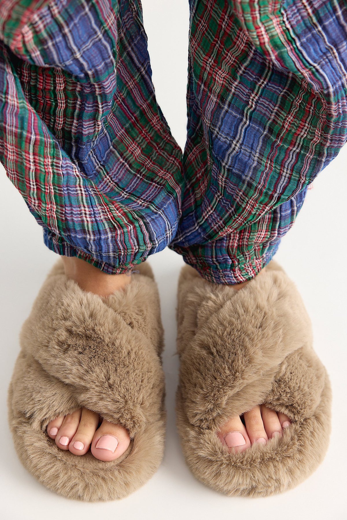 A close-up of a person wearing fluffy taupe-brown slippers with their feet visible, paired with blue and green plaid pajama pants, against a plain white background.