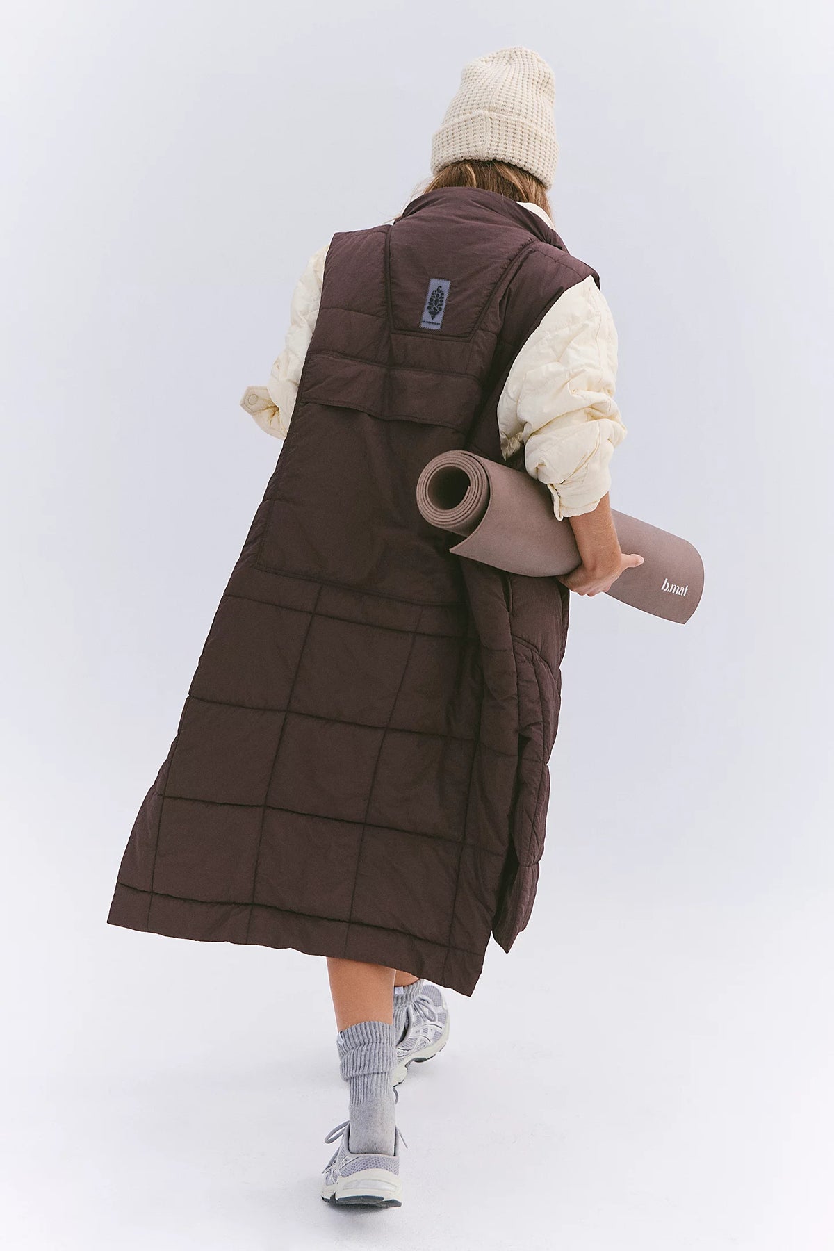 A person shown from behind wearing a long dark brown quilted puffer vest over a cream long-sleeve top, with a cream beanie, gray socks, and white sneakers. The person is holding a rolled-up taupe yoga mat against their back, set against a clean white studio background.