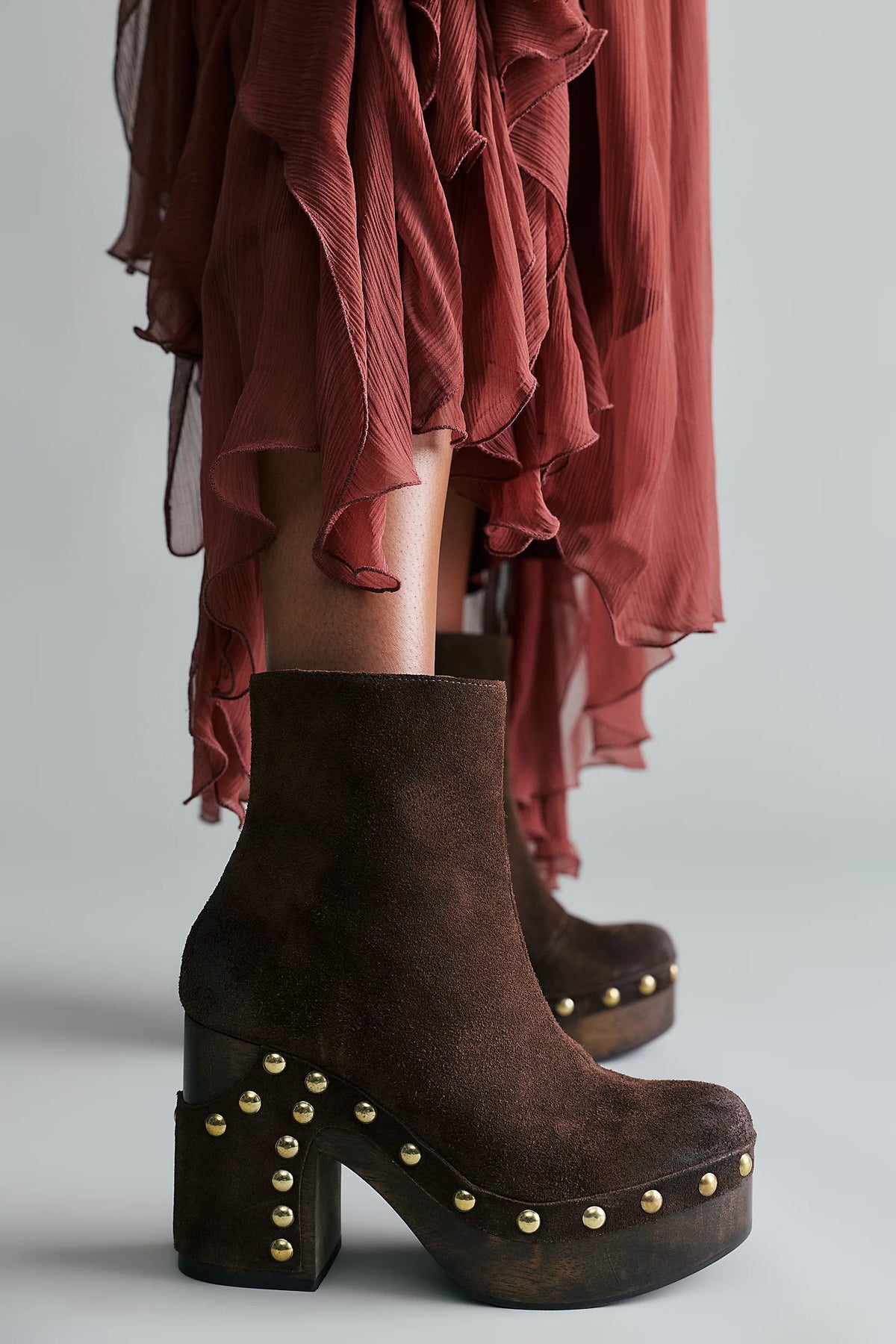 A close-up of a person wearing dark brown suede platform ankle boots with gold-tone studs, paired with a rust-colored sheer ruffled skirt, against a light gray studio background.