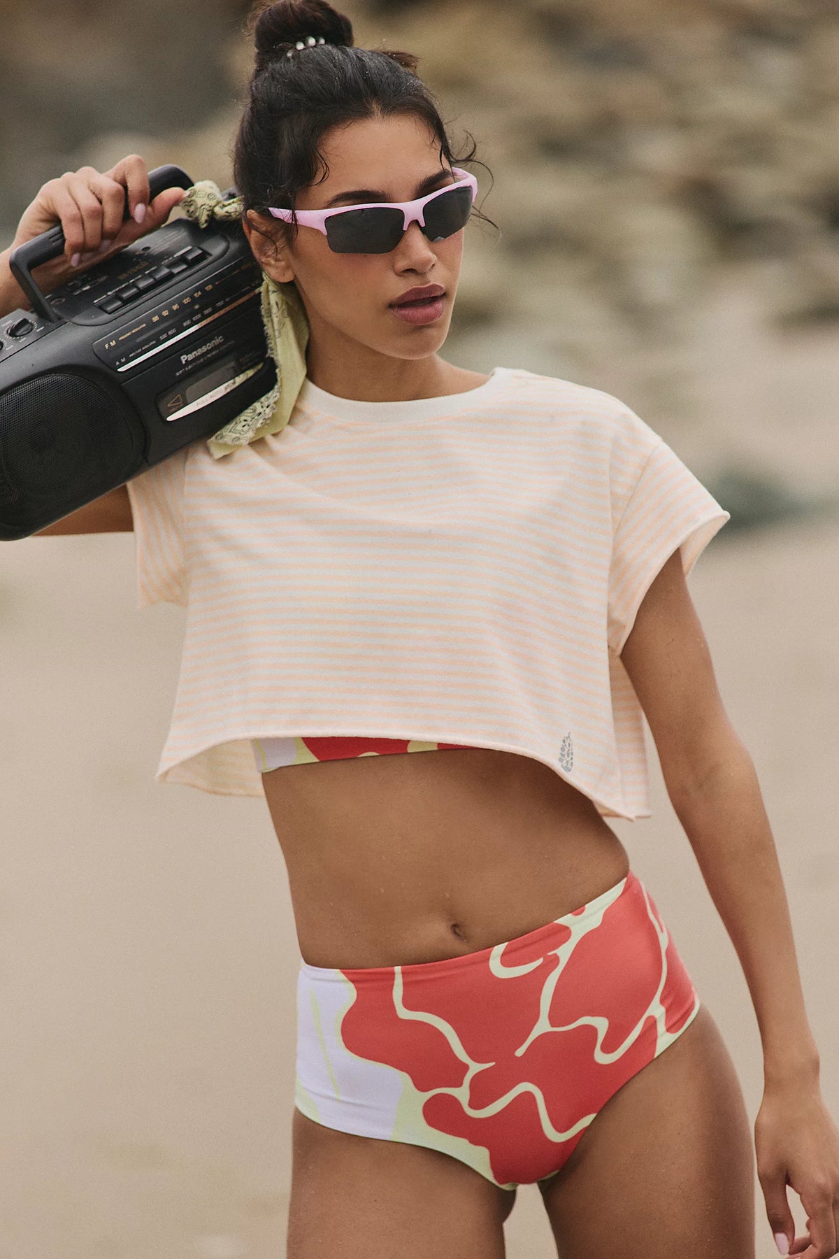 Woman in a white crop top and colorful shorts holding a boombox on a beach.