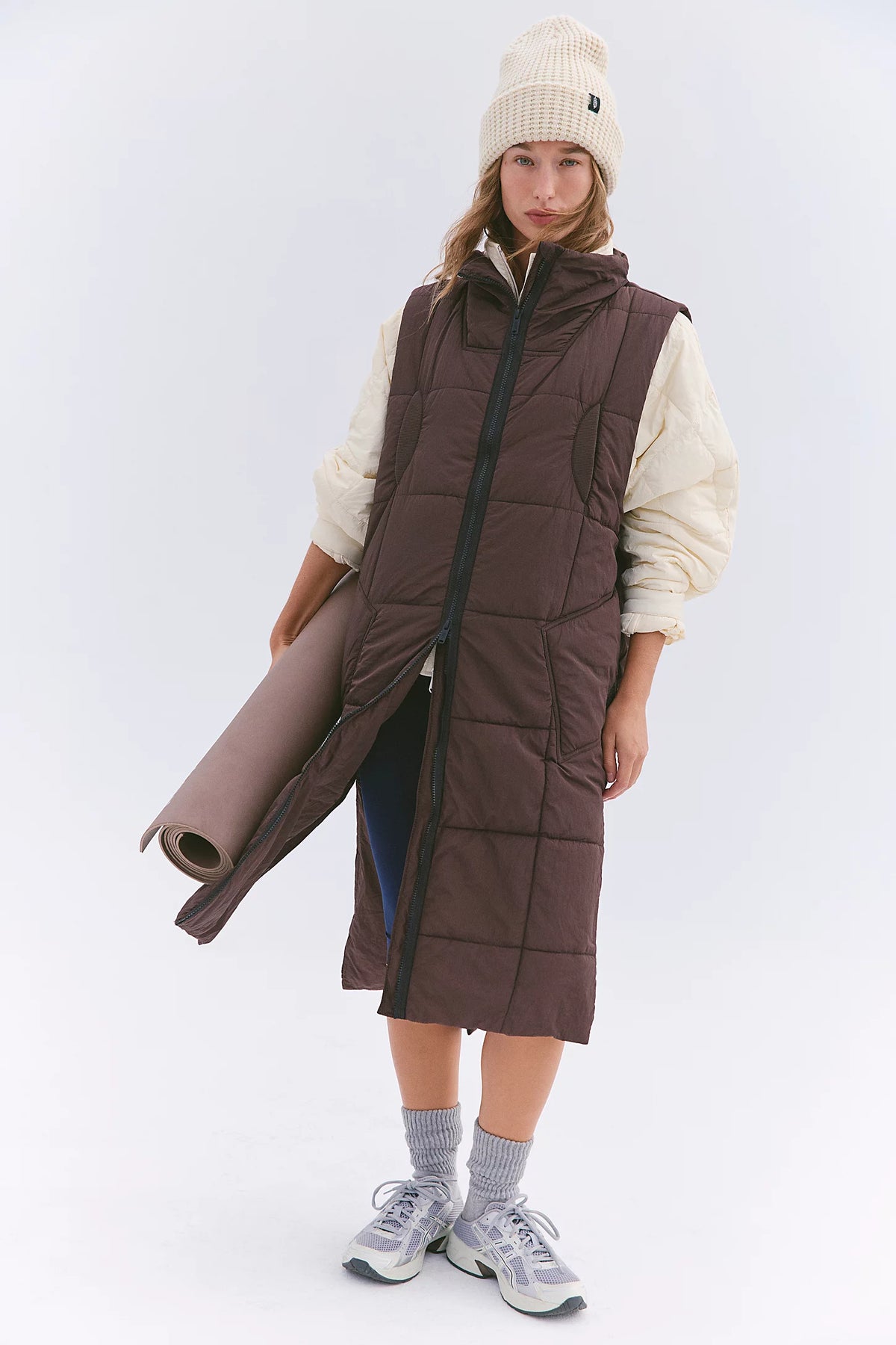 A model wearing a long brown quilted puffer vest over a cream-colored long-sleeve top, paired with a cream beanie, gray socks, and white sneakers, holding a rolled-up brown mat against a light gray studio background.