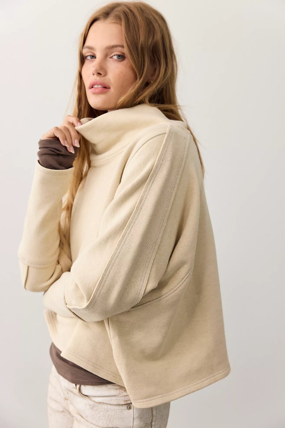 A woman wearing a cream-colored oversized cropped hoodie with a high mock neck, layered over a brown long-sleeve shirt and light beige pants, photographed against a plain light gray studio background.