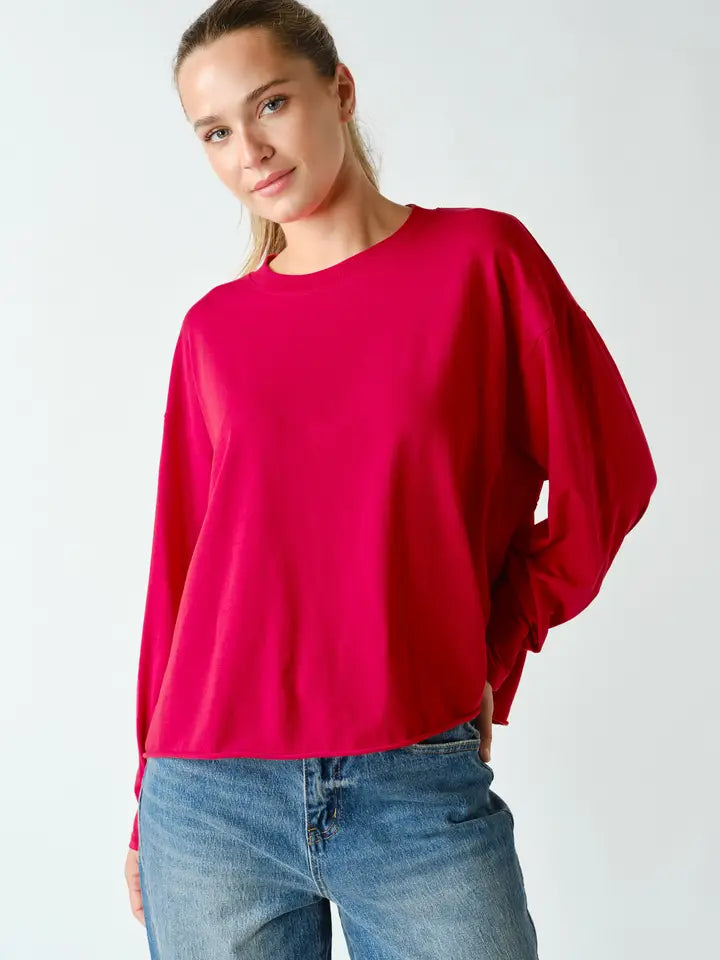 A woman wearing a bright fuchsia pink long-sleeve crewneck top and blue jeans, photographed against a clean light gray studio background.