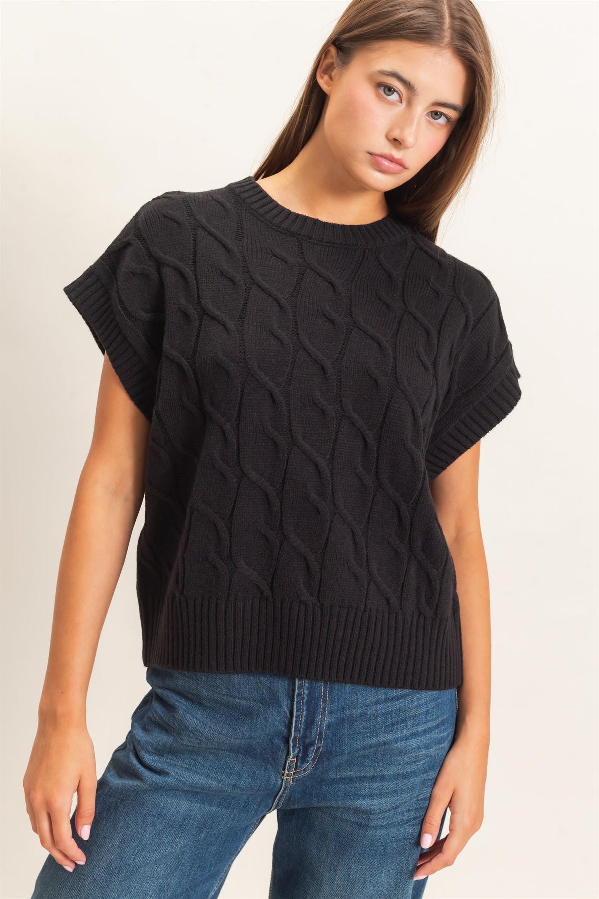 A woman wearing a black short-sleeve cable-knit sweater and blue jeans, photographed against a plain light background.