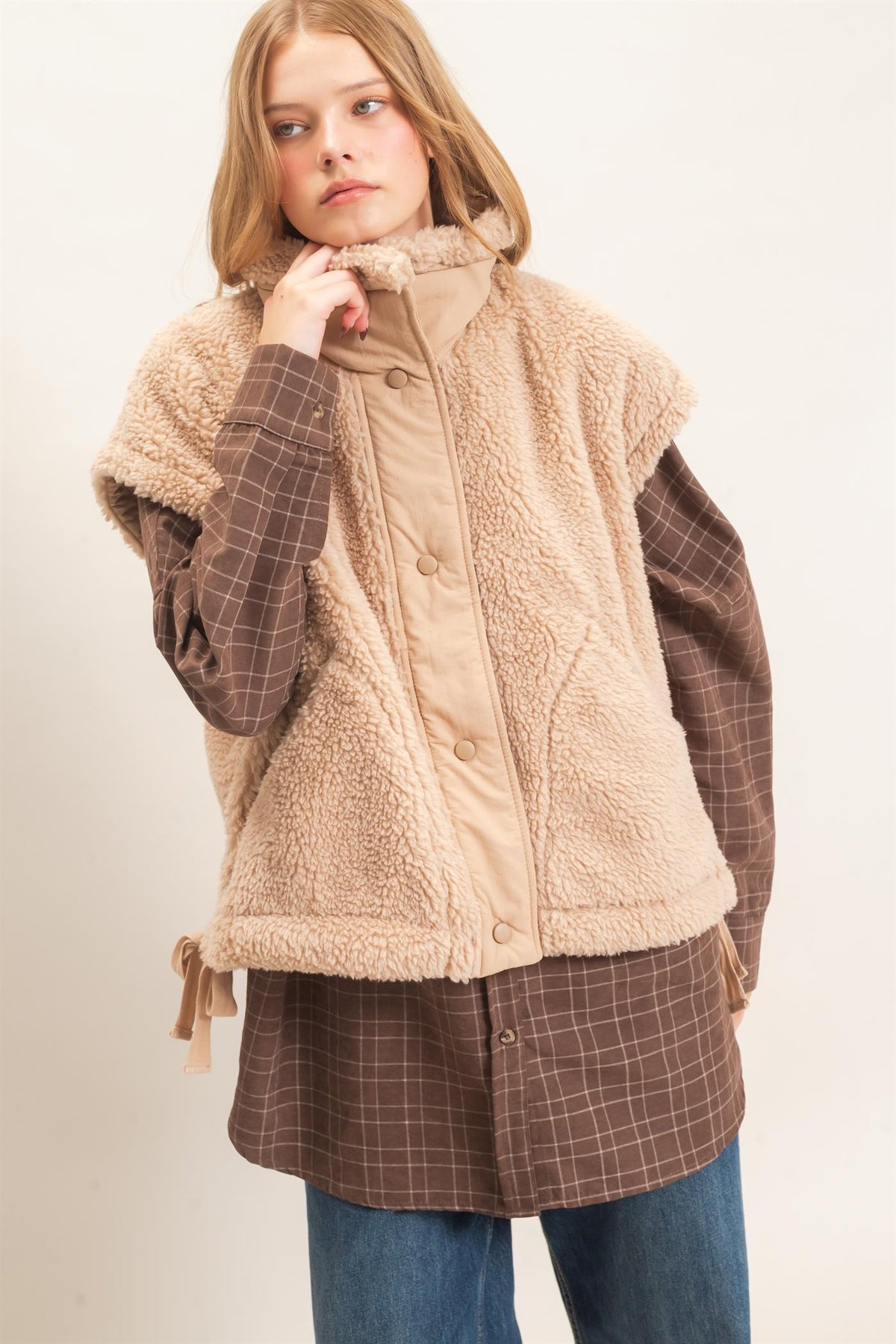 A model wearing a beige sherpa fleece vest over a brown plaid button-up shirt and blue jeans, photographed against a plain off-white studio background.