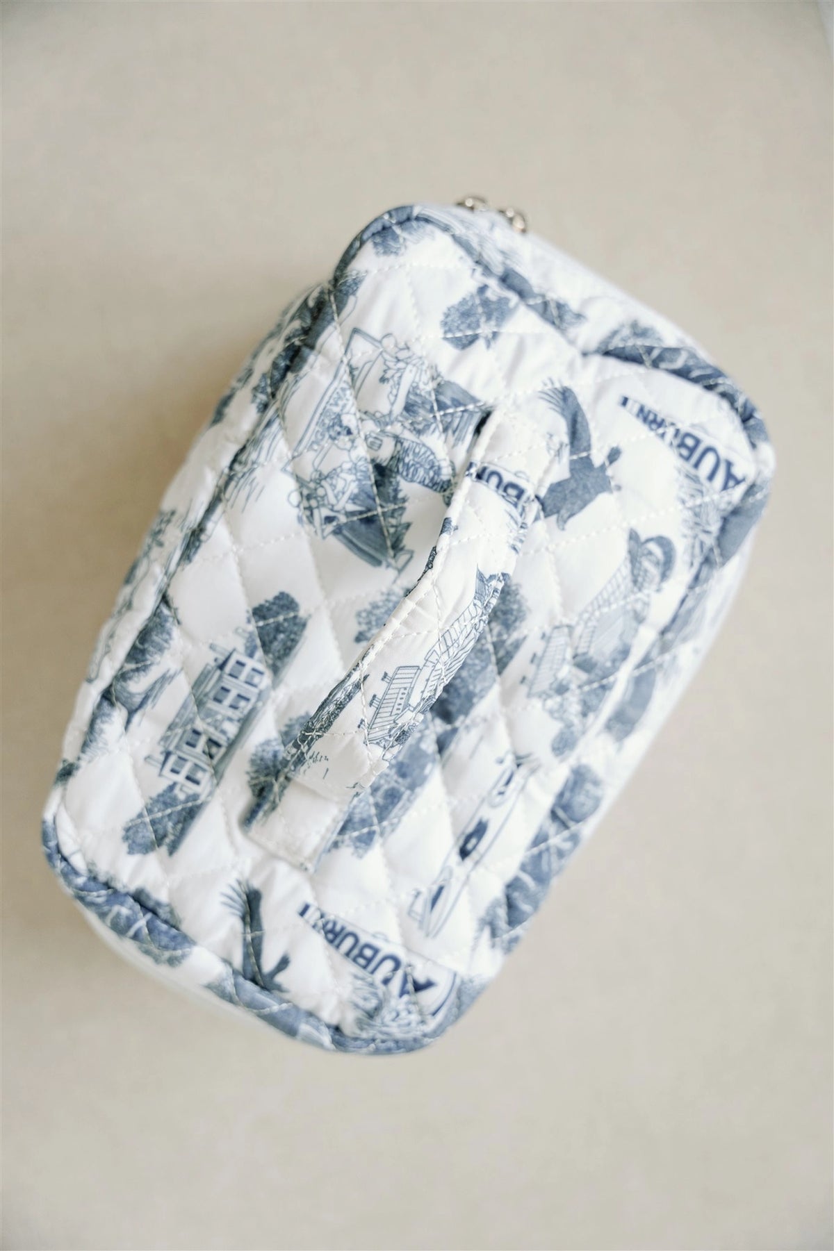 A quilted cosmetic pouch with a blue and white toile de Jouy pattern featuring the word 'AUBURN' printed repeatedly, shown on a neutral beige background.