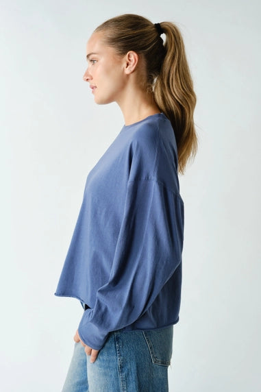 A woman in profile wearing a loose, oversized blue long-sleeve top and blue jeans against a plain light gray studio background.