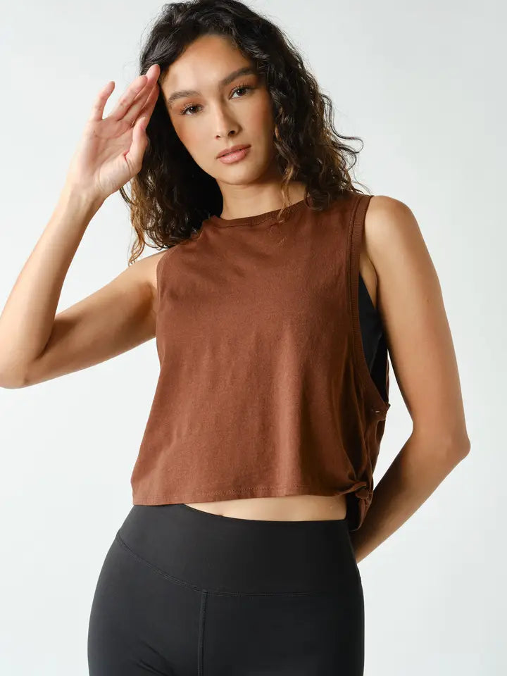 A woman modeling a sleeveless brown crop top with a side tie detail, paired with black leggings, against a light gray studio background.