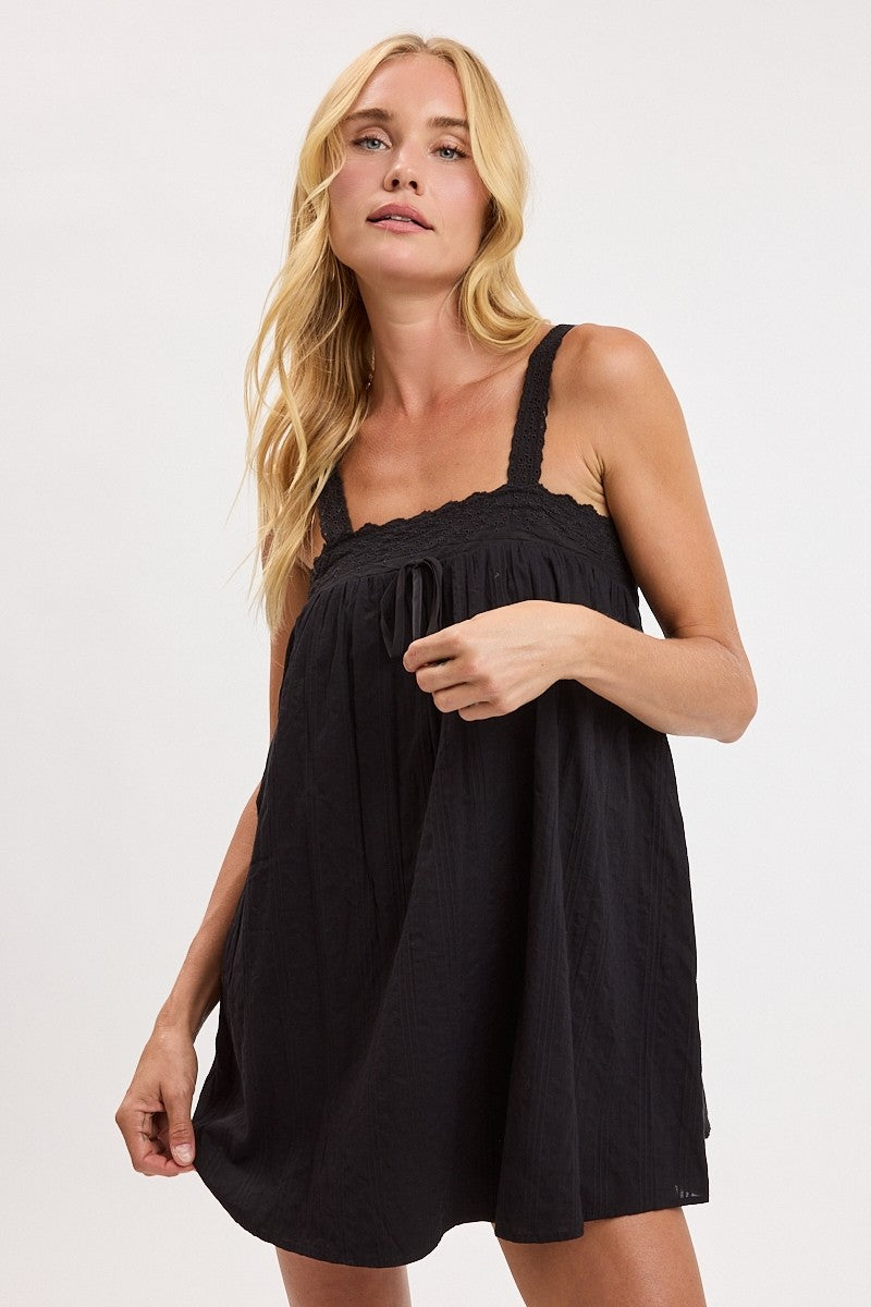 A blonde woman wearing a black sleeveless mini dress with a gathered smocked bodice and wide shoulder straps, photographed against a clean white studio background.