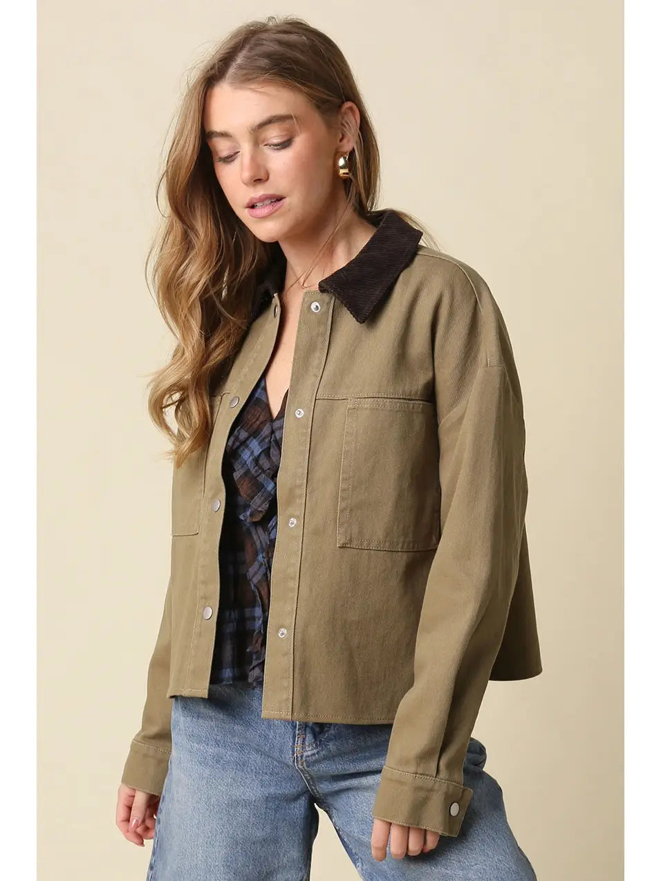 A woman wearing an olive green utility jacket with a dark brown corduroy collar, layered over a plaid shirt and blue jeans, set against a solid beige background.