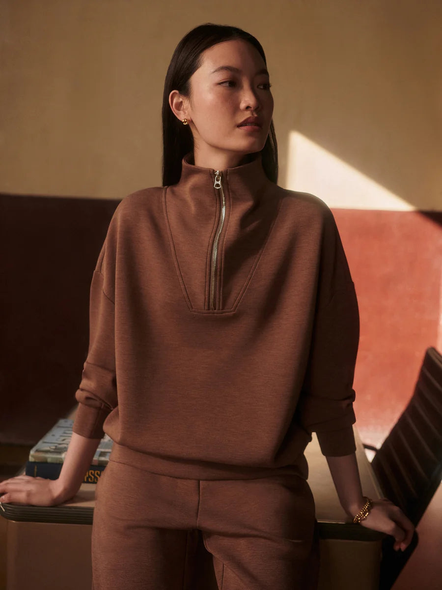 A woman wearing a matching brown knit lounge set with a half-zip mock neck sweatshirt and coordinating pants, posed in a warm-toned indoor setting.