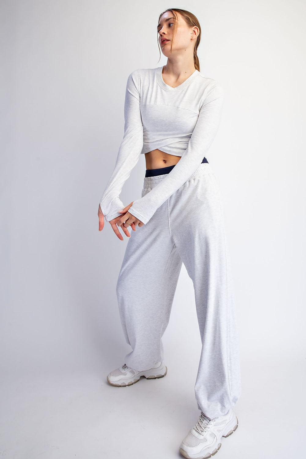 A woman modeling a white long-sleeve crop top and matching white sweatpants with white sneakers, standing against a light gray studio background.