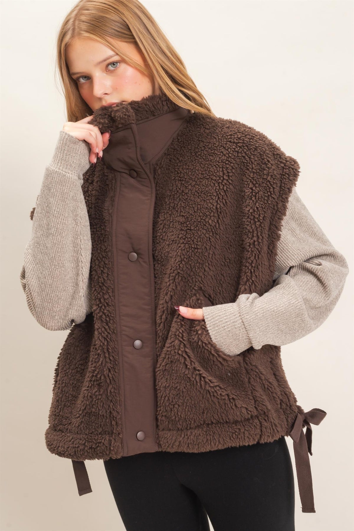 A woman wearing a brown sherpa fleece vest over a light gray knit sweater and black pants, photographed against a plain light beige studio background.