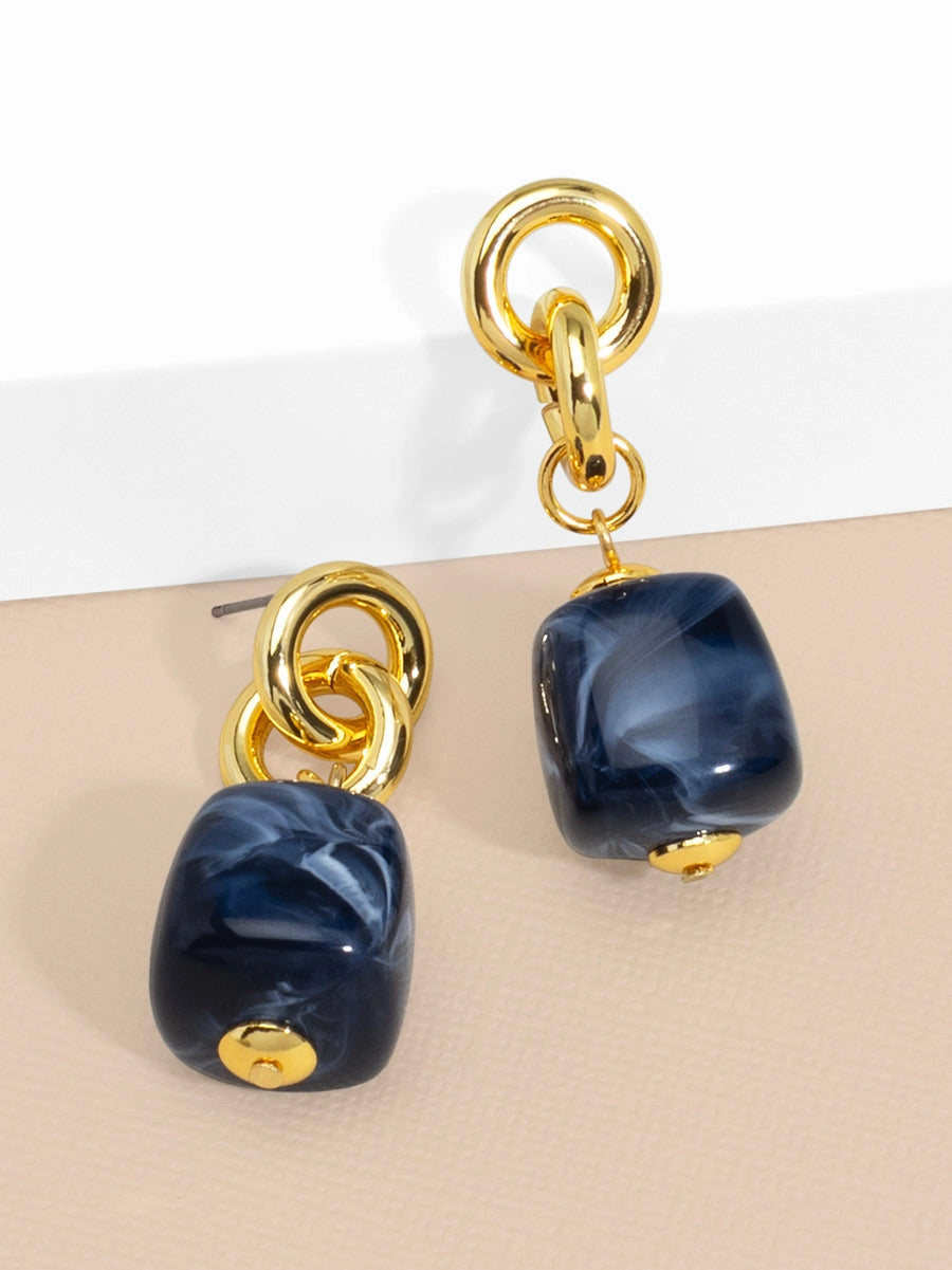 A pair of gold-tone drop earrings featuring a polished, rounded dark blue marbled bead suspended from two interlocking gold rings, displayed on a beige surface with a white background.