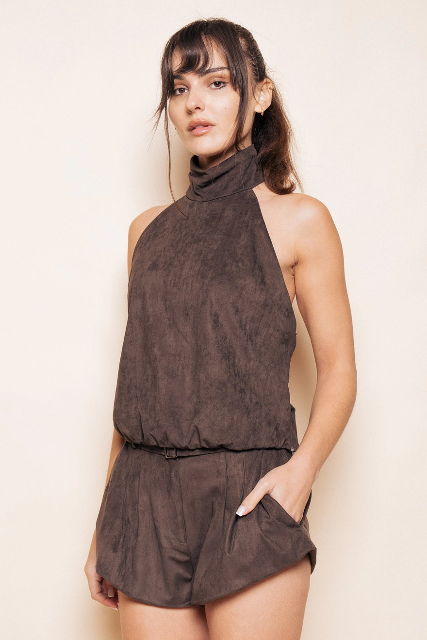 Model with brown hair wearing a brown suede halter tank top with matching brown bubble shorts with pockets standing in front of a beige background. 