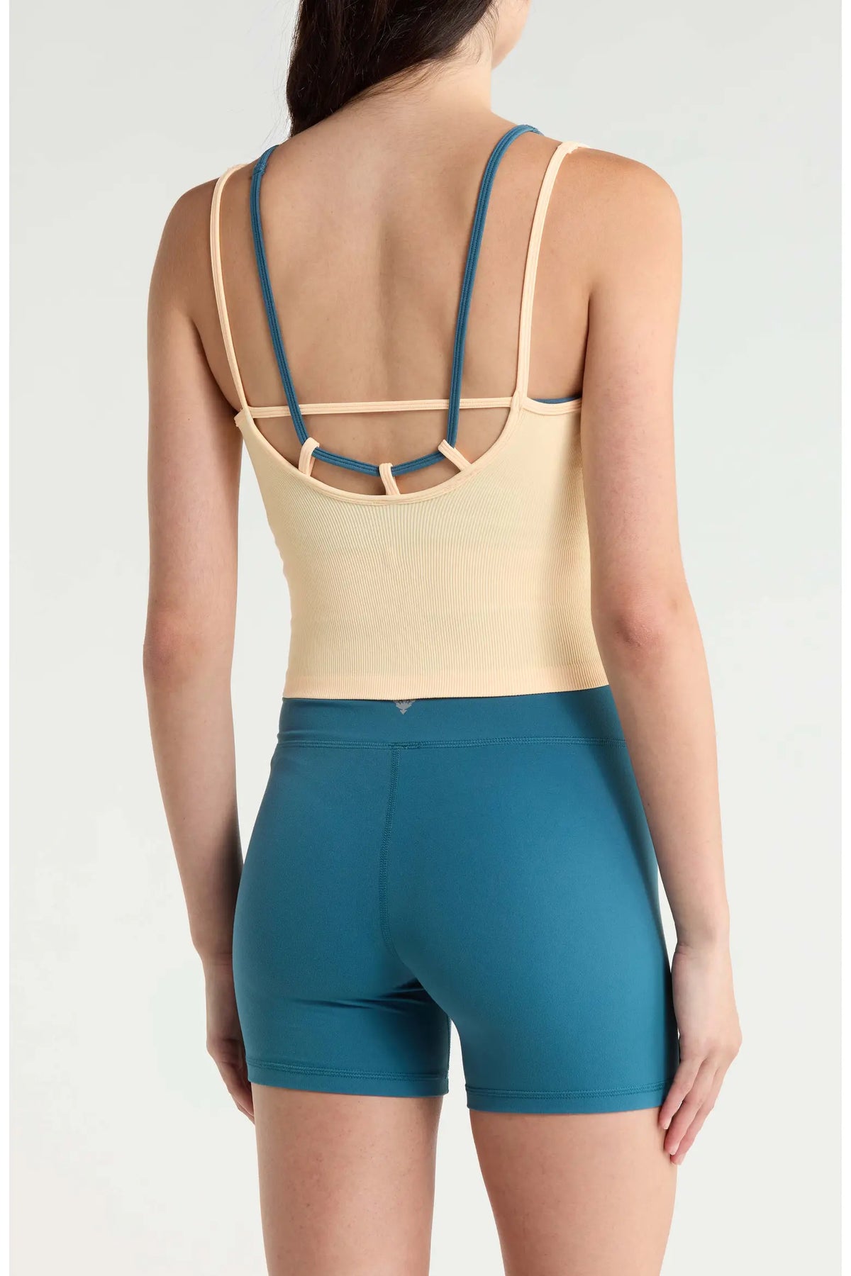 A woman shown from the back wearing a beige strappy bodysuit with teal trim and matching teal athletic shorts against a light gray studio background.