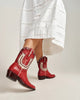 Red leather cowboy boots with white western embroidery, shown on a model wearing a white skirt against a light gray studio background.