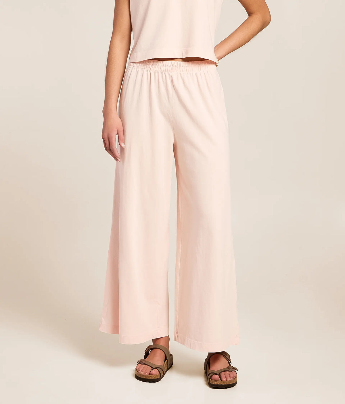 A model wearing light pink wide-leg cropped pants with an elastic waistband, paired with brown strappy sandals, against a solid beige studio background.