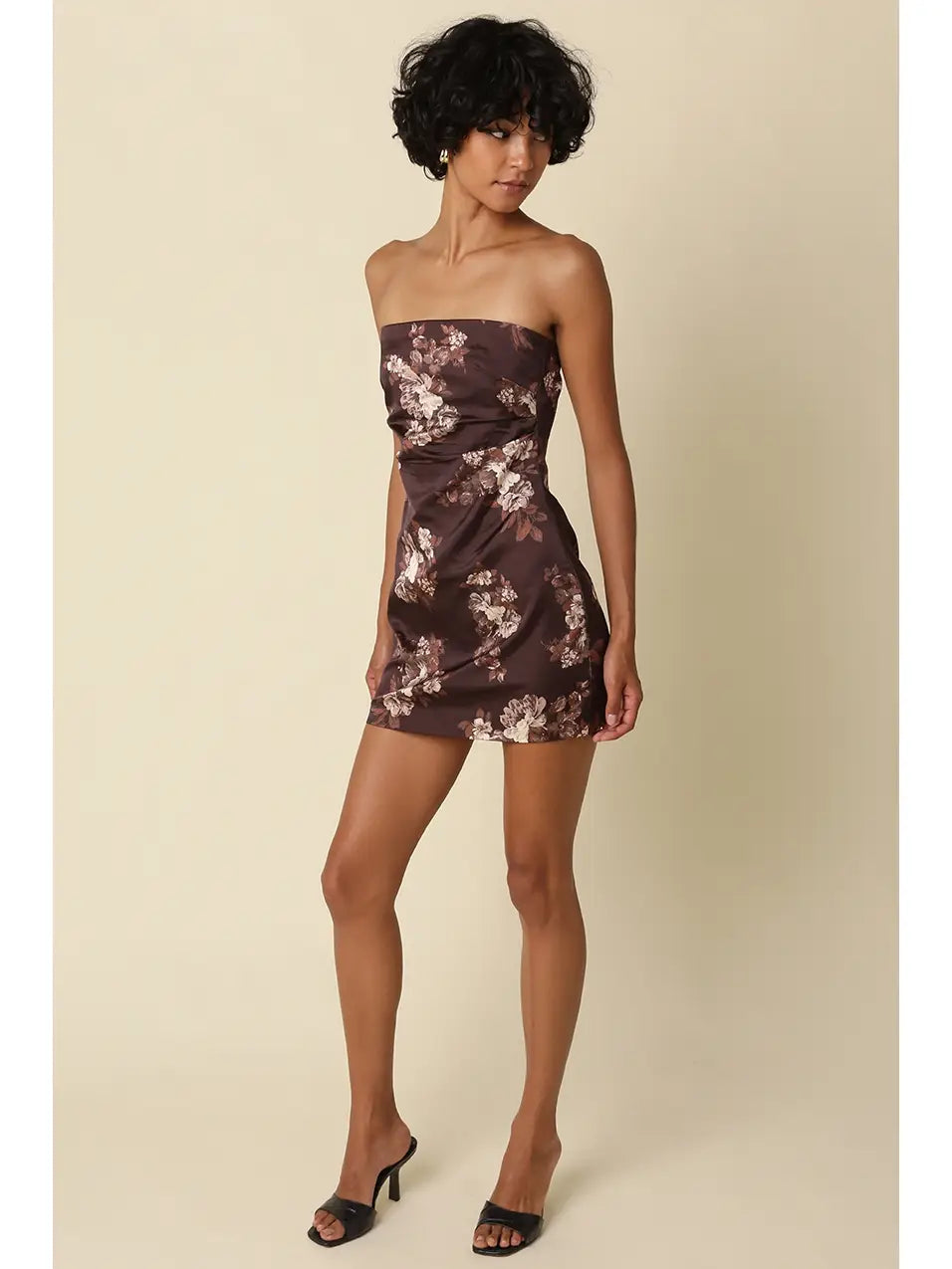 A woman modeling a strapless mini dress with a brown and beige floral pattern, paired with black heeled sandals, against a solid beige background.