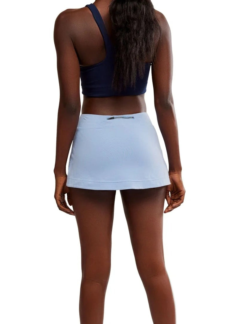 A woman shown from the back wearing a light blue athletic tennis skirt, a navy blue sports bra, and black athletic sneakers against a solid white background.