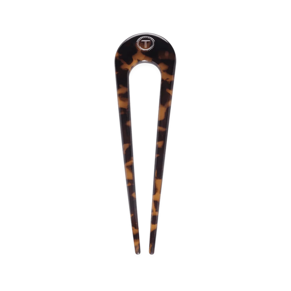 A single tortoiseshell hair divot tool with a small circular T-shaped logo at the top, shown vertically on a plain white background.