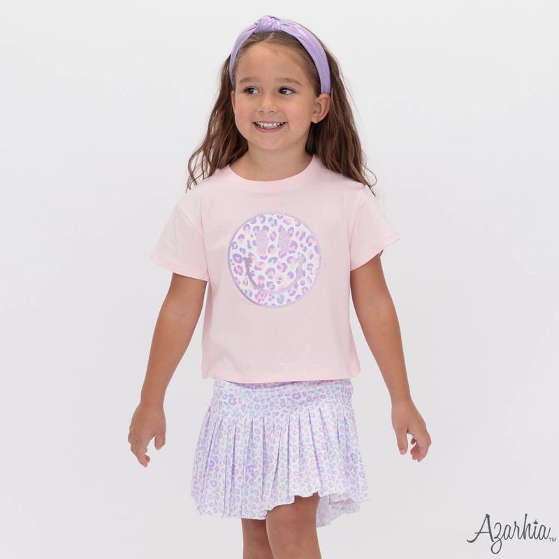 A young girl smiling and wearing a light pink short-sleeve t-shirt with a purple and white patterned circular graphic on the chest, paired with a matching light purple patterned ruffled skirt and a lavender headband. The brand name 'Agarkia.