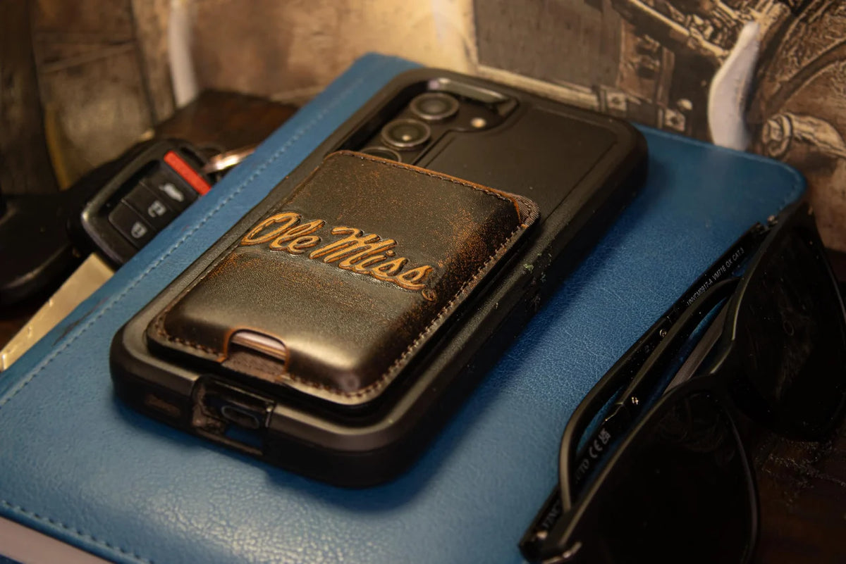 A black rugged phone case with a brown faux-leather wallet insert featuring the words 'Ole Miss' in gold script, resting on a blue leather folio next to sunglasses.