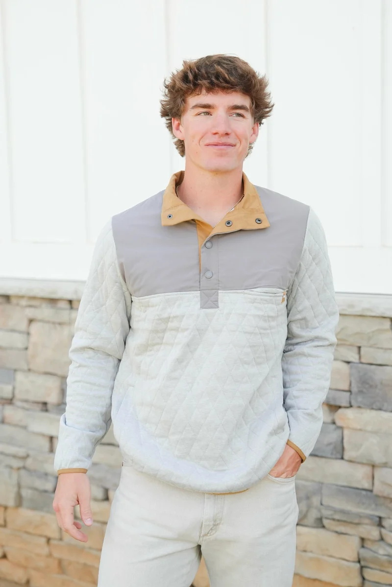 A man wearing a color-blocked long-sleeve pullover with a light gray quilted body, a tan collar, and a gray yoke, standing in front of a stone wall.