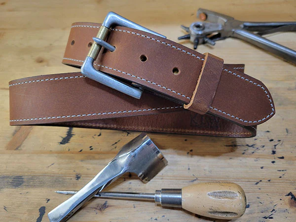 Brown leather belt with silver buckle displayed on wooden workbench with leather tools.