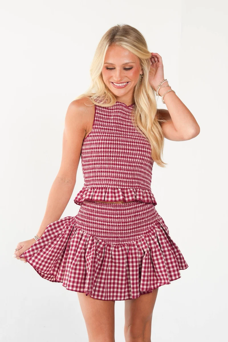 A blonde woman modeling a sleeveless red and white gingham mini dress with a smocked waist and ruffled skirt, set against a plain white studio background.