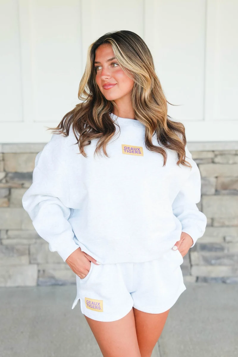 Woman wearing a white sweatshirt and shorts with logos, standing against a stone wall.