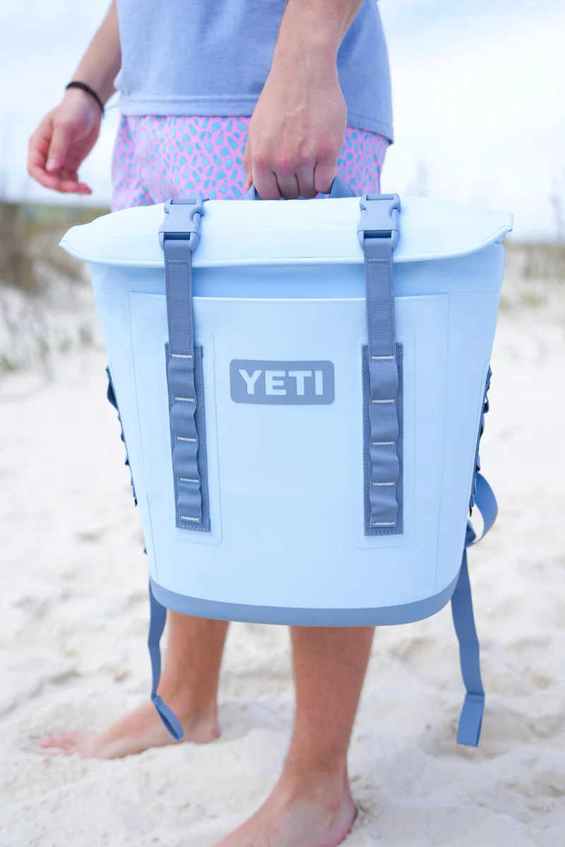 Person holding a light blue YETI soft cooler bag while standing barefoot on sandy beach.