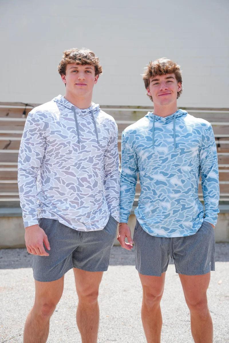 Two young men standing outdoors wearing matching long-sleeve hooded shirts with a blue and white abstract pattern, paired with gray shorts.