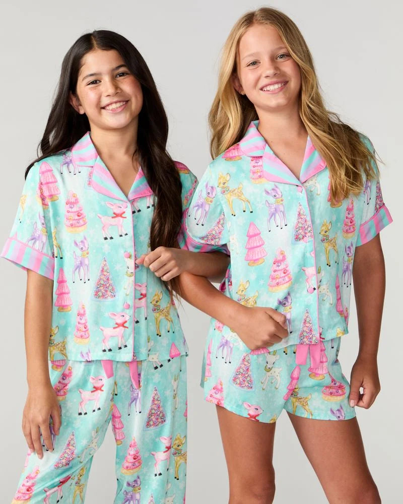 Two girls modeling matching short-sleeve pajama sets with a pastel unicorn and castle print on a mint green background, against a light gray studio backdrop.
