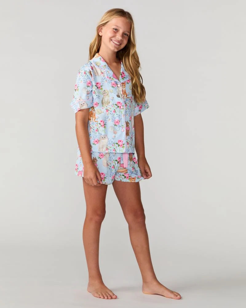 A young girl modeling a light blue short-sleeve pajama set with a colorful floral and bunny print, standing against a neutral light gray background.