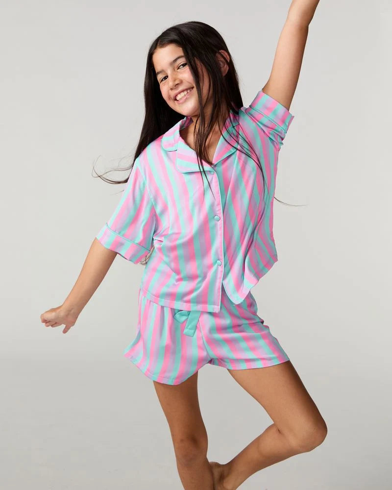 A young girl wearing a pink and mint green striped pajama set, including a short-sleeve button-up top and matching shorts, posing dynamically against a light gray studio background.