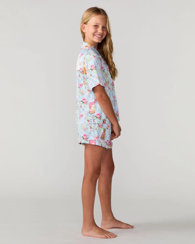 A young girl with long blonde hair stands in profile against a light gray studio background, wearing a short-sleeve button-front shirt and matching shorts set with a colorful floral and bunny print on a pale blue base.