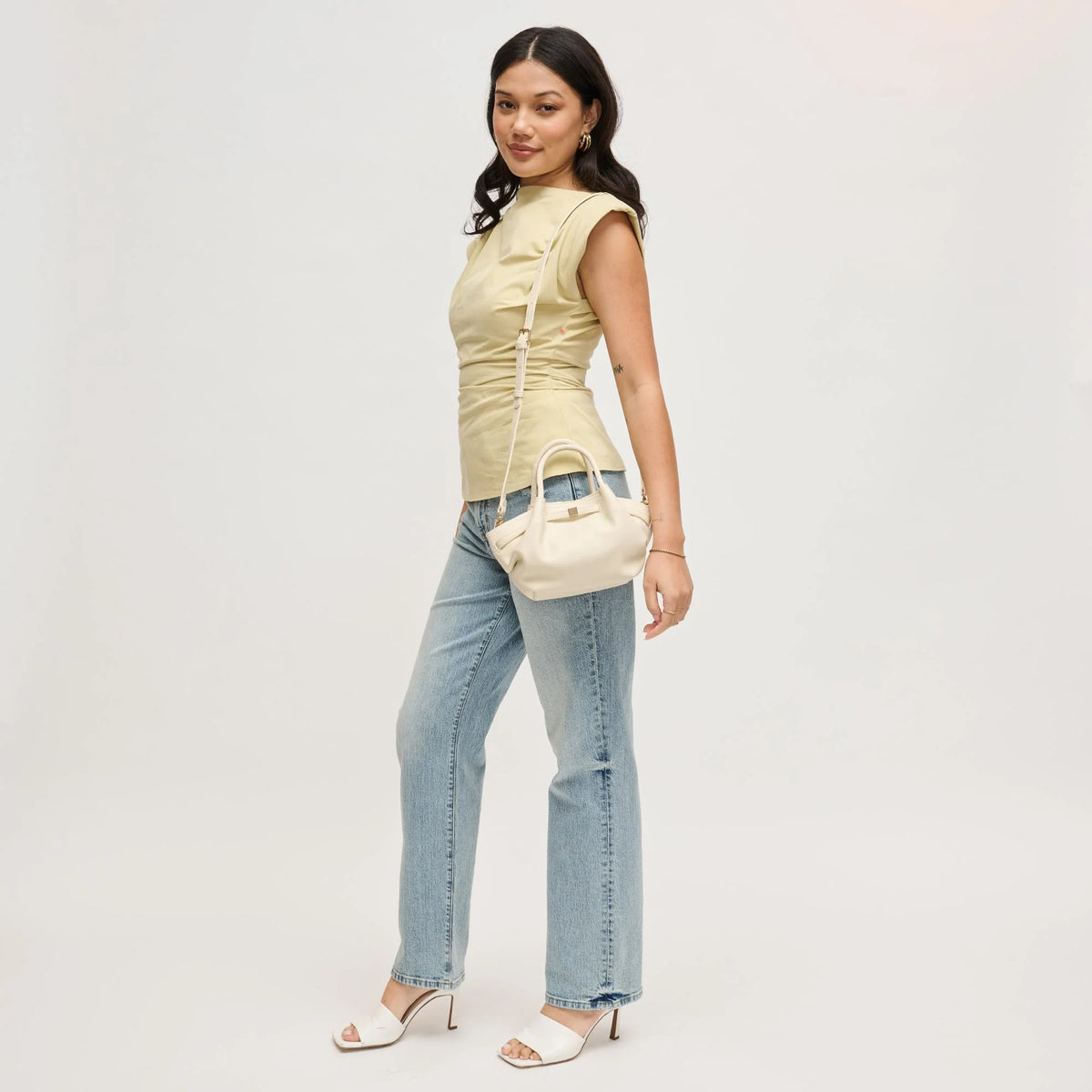 Woman wearing a light-colored sleeveless top and blue jeans, holding a cream-colored handbag on a plain background