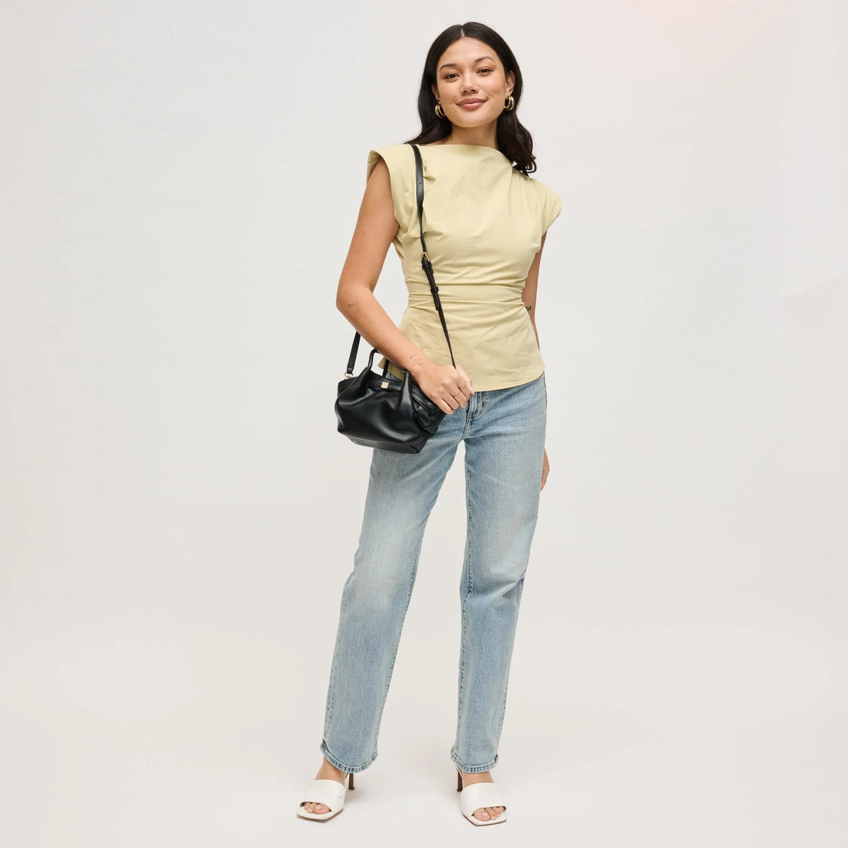 Woman wearing a light yellow top and blue jeans, holding a black handbag on a white background