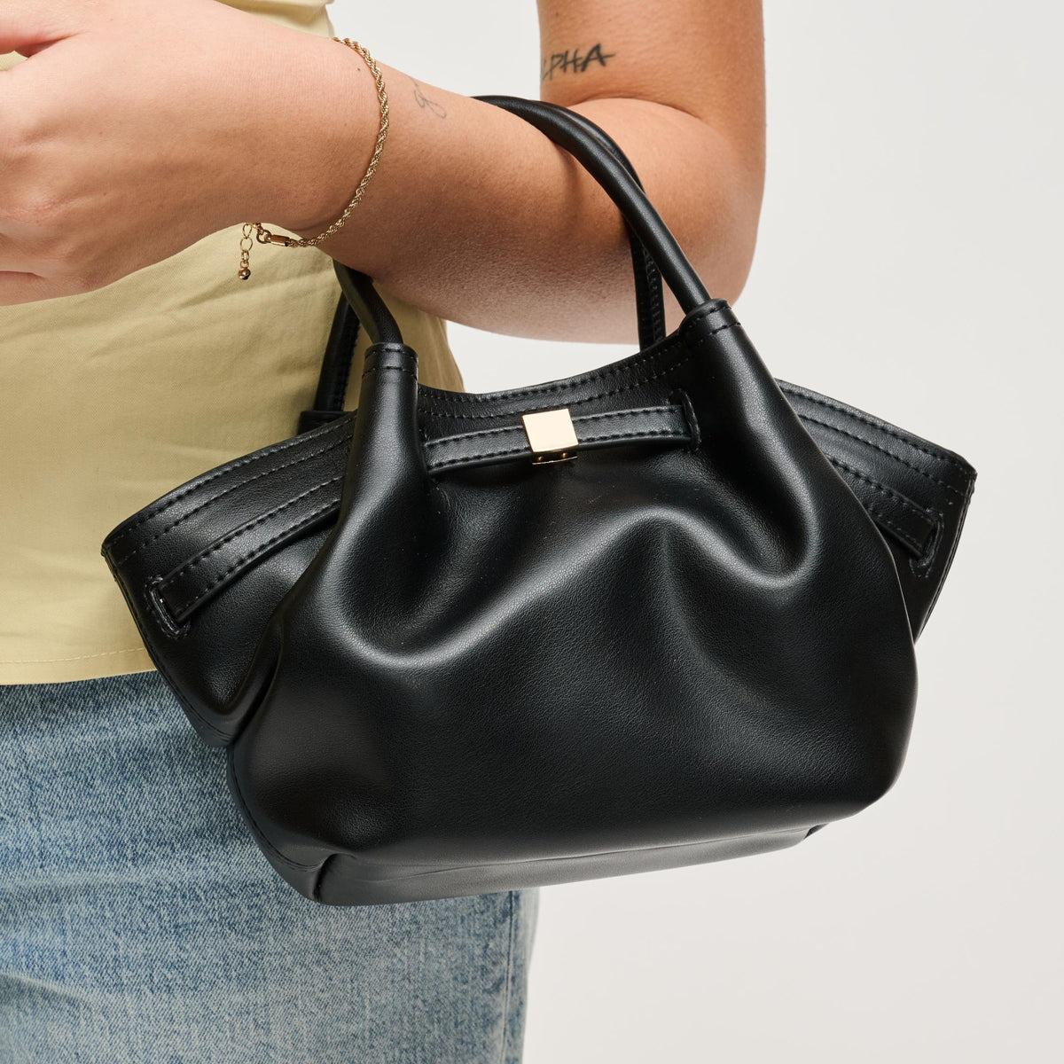 Black handbag held by a person wearing a beige top and blue jeans on a light gray background
